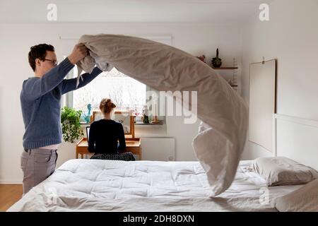 Man making bed while woman working at table in bedroom Stock Photo