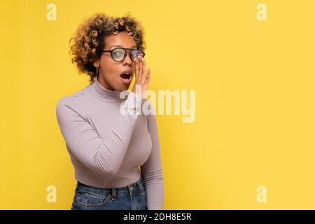 Young african american woman with hand on mouth telling secret rumor. Studio shot on yellow wall. Stock Photo