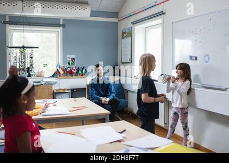 Teacher and students looking at girls discussing while standing against whiteboard Stock Photo