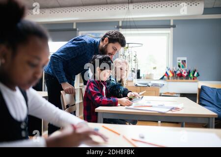 Teacher looking at boy and girl using digital tablet in classroom Stock Photo
