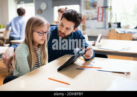 Teacher discussing with student while showing digital tablet at desk in classroom Stock Photo