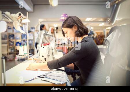 Side view of woman sewing fabric on machine while volunteers working in workshop Stock Photo