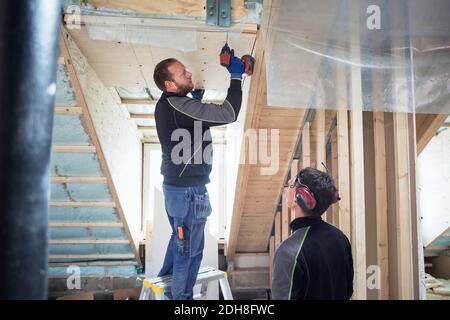 Coworker looking at carpenter drilling on roof beam in construction site Stock Photo