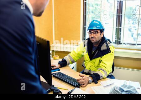 Serious construction worker in protective wear talking to man at desk Stock Photo