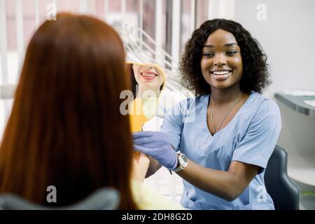 Back view of woman patient looking at her smile after curing or whitening teeth in dental clinic, and happy African female dentist, showing the mirror Stock Photo