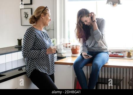 Mother talking with happy daughter using smart phone in kitchen Stock Photo