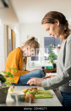 Woman cutting fruits with girl sitting on kitchen counter at home Stock Photo