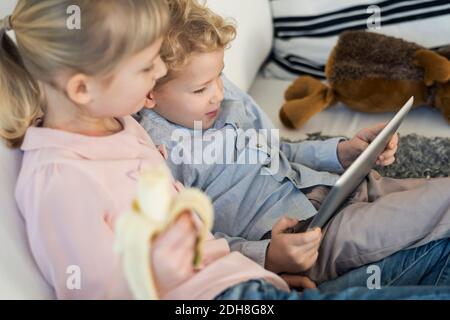 Siblings using digital tablet while eating banana at home Stock Photo