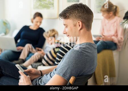 Side view of man using phone with family sitting on sofa at home Stock Photo