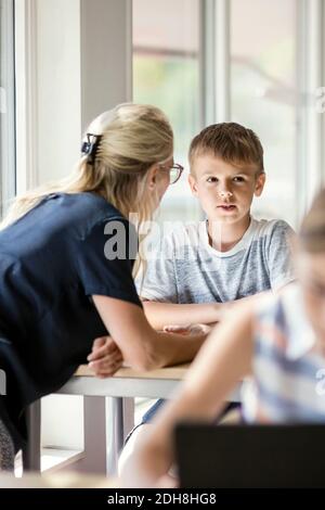Teacher assisting boy at desk with girl in foreground
