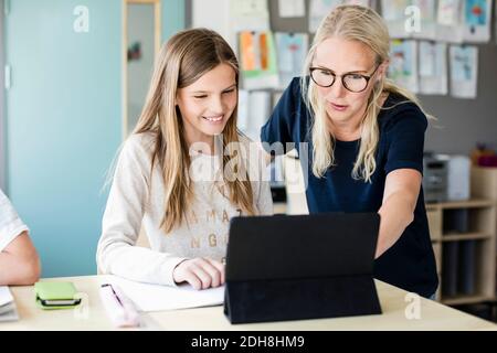 Teacher assisting happy girl in using digital tablet at school Stock Photo