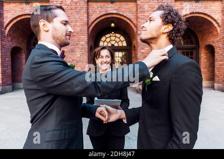 Man adjusting bow tie of gay partner in front of priest during wedding ceremony Stock Photo