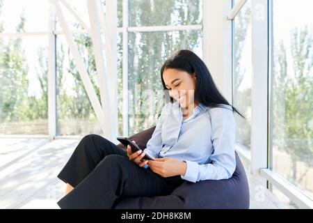 Beautiful young african woman looking at smart phone and smiling while sitting on bean bag chair with panoramic windows backgrou Stock Photo