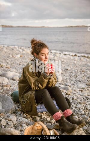 Thoughtful woman sitting on rock with drink at beach Stock Photo