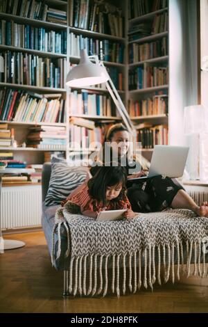 Mother using laptop while down syndrome daughter with digital tablet on sofa in living room Stock Photo