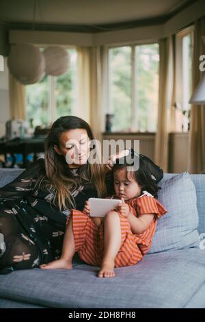 Mother looking at down syndrome daughter using mobile phone while sitting on sofa in living room Stock Photo