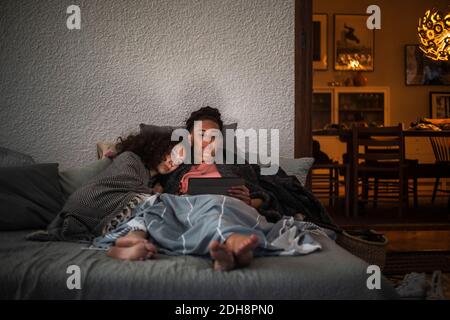 Mother and daughter watching digital tablet together while relaxing on sofa at home Stock Photo