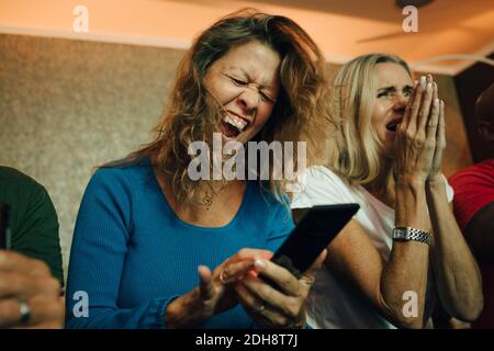 Happy woman cheering by friends while watching sports on smart phone in living room Stock Photo