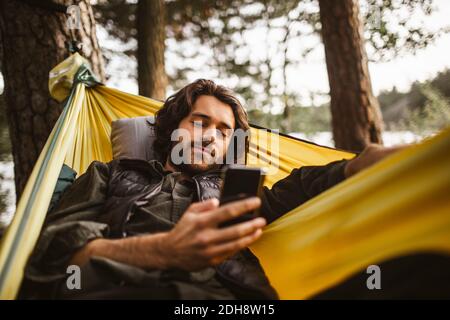Young man using smart phone while lying over hammock in forest Stock Photo