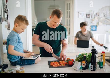 Father cutting tomato while son using digital tablet on kitchen counter Stock Photo