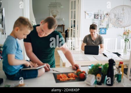 Man using laptop dining table while father and son looking at digital tablet on kitchen counter Stock Photo