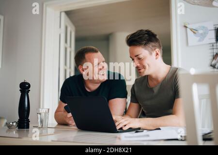 Male talking to smiling partner while using laptop over dining table at home Stock Photo
