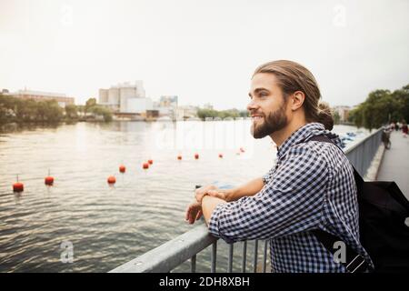 Thoughtful smiling man standing at railing by river in city against clear sky Stock Photo