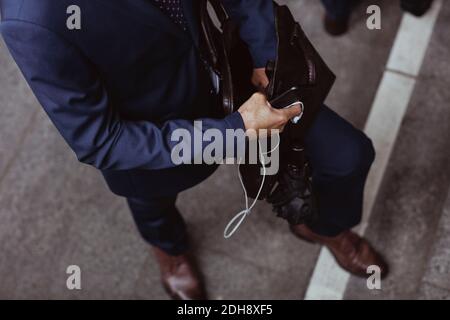 Low section of businessman with open bag holding smart phone and in-ear headphones in city Stock Photo