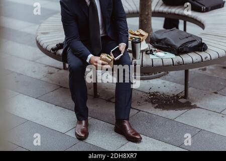 Low section of entrepreneur with smart phone and sandwich sitting in city Stock Photo