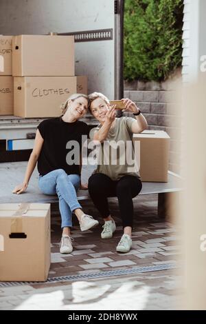 Female friends taking selfie while sitting cardboard boxes during relocation Stock Photo