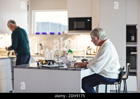 Senior woman using laptop while male preparing food in kitchen Stock Photo
