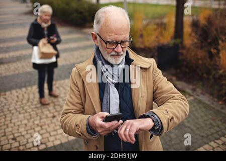 Senior man with smart phone checking the time while partner standing behind on footpath Stock Photo