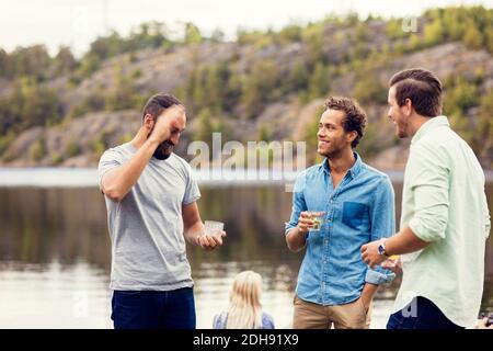 Friends talking and having drink while standing at lakeshore Stock Photo