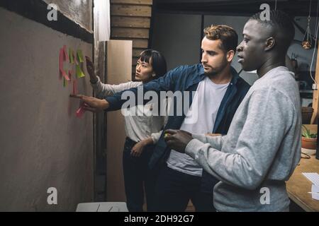 Male and female coworkers sticking adhesive note on wall at workplace Stock Photo