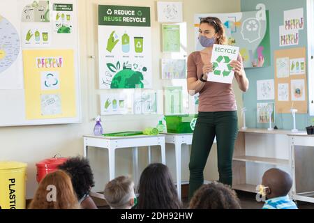 Female teacher wearing face mask showing recycle symbol to students in class Stock Photo