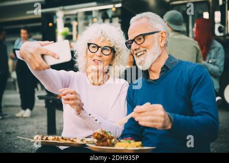 Senior woman taking selfie with smiling man eating meal against food truck in city Stock Photo
