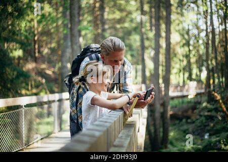Smiling father with backpack and daughter looking at smart phone in forest Stock Photo