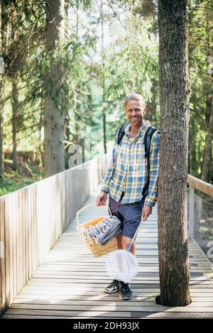 Portrait of smiling man with picnic basket standing on footbridge in forest Stock Photo