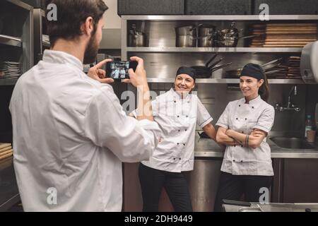 Male chef taking photograph of smiling female coworkers in commercial kitchen Stock Photo