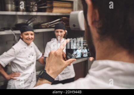 Male chef taking photograph of female coworkers in commercial kitchen Stock Photo