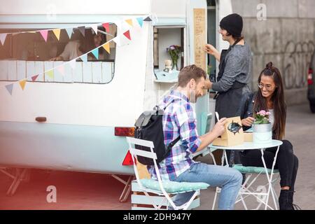 Happy couple sitting at table outside food truck while owners working in background Stock Photo
