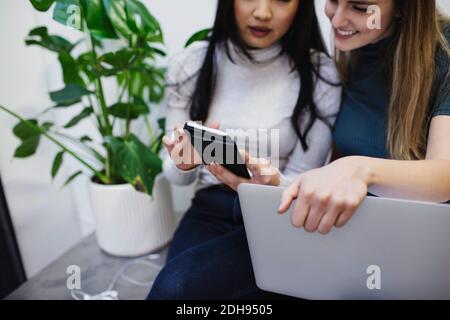 Midsection of female bloggers using smart phone while sitting on sideboard in creative office Stock Photo