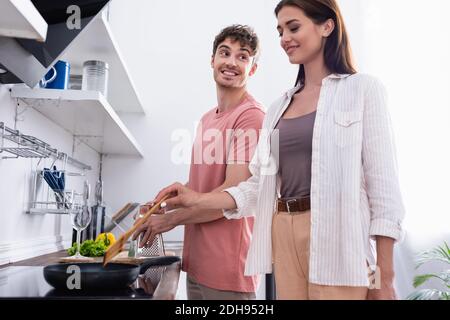 Smiling man looking at girlfriend cooking on stove in kitchen Stock Photo