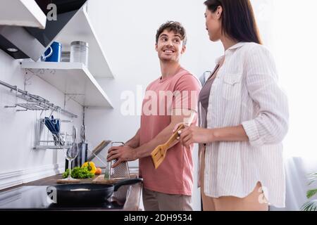 Smiling man holding grater near vegetables and girlfriend with spatula near stove on blurred foreground Stock Photo