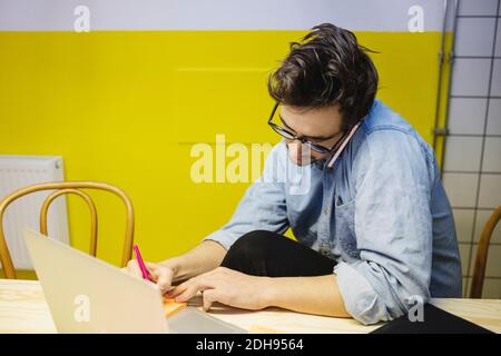 Young man using smart phone while writing on adhesive note at desk in board room Stock Photo