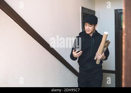Smiling delivery man with package using phone against wall Stock Photo