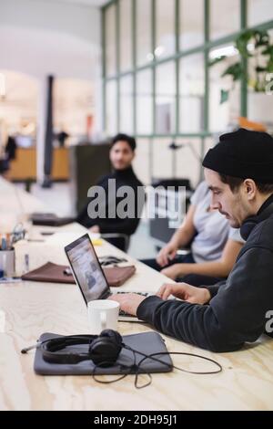 Young man using laptop at desk while colleagues sitting in background Stock Photo