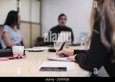 Midsection of young woman writing at desk while colleagues sitting in background Stock Photo