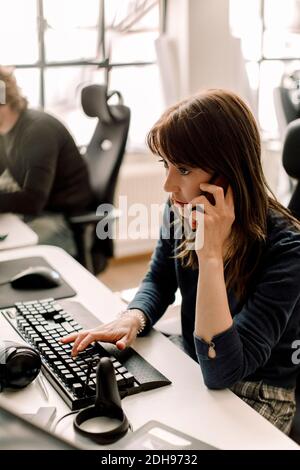 Female entrepreneur working on programming while talking on phone at workplace Stock Photo