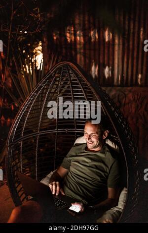 Smiling man using laptop while sitting in hanging chair at night Stock Photo
